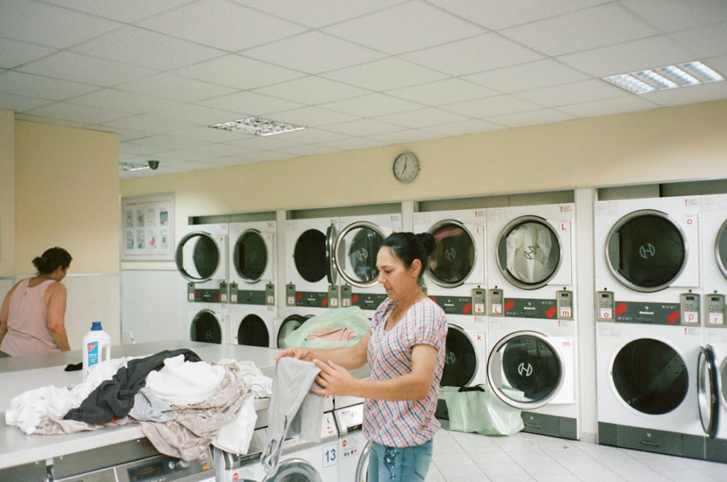 A woman folds clothes inside a laundromat lined with rows of washing machines and dryers.