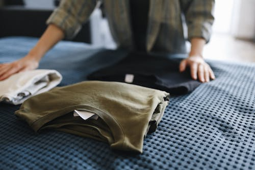 A person arranges neatly folded clothes on a bed, preparing or organizing outfits.